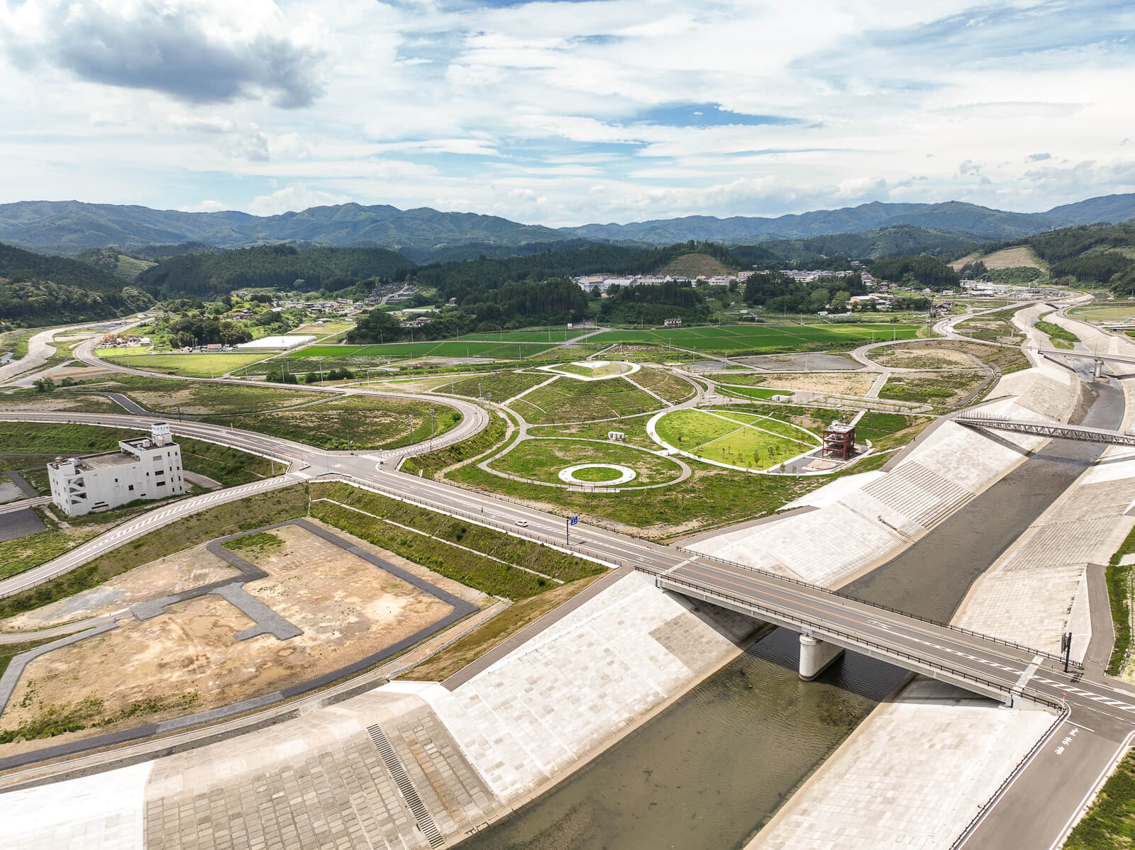 奥に向かって緩やかに蛇行する川の堤防にかかる橋と、その周囲の整地された土地や道路、遠景に山々と緑地が広がる昼間の風景を高台から定点カメラで撮影した写真