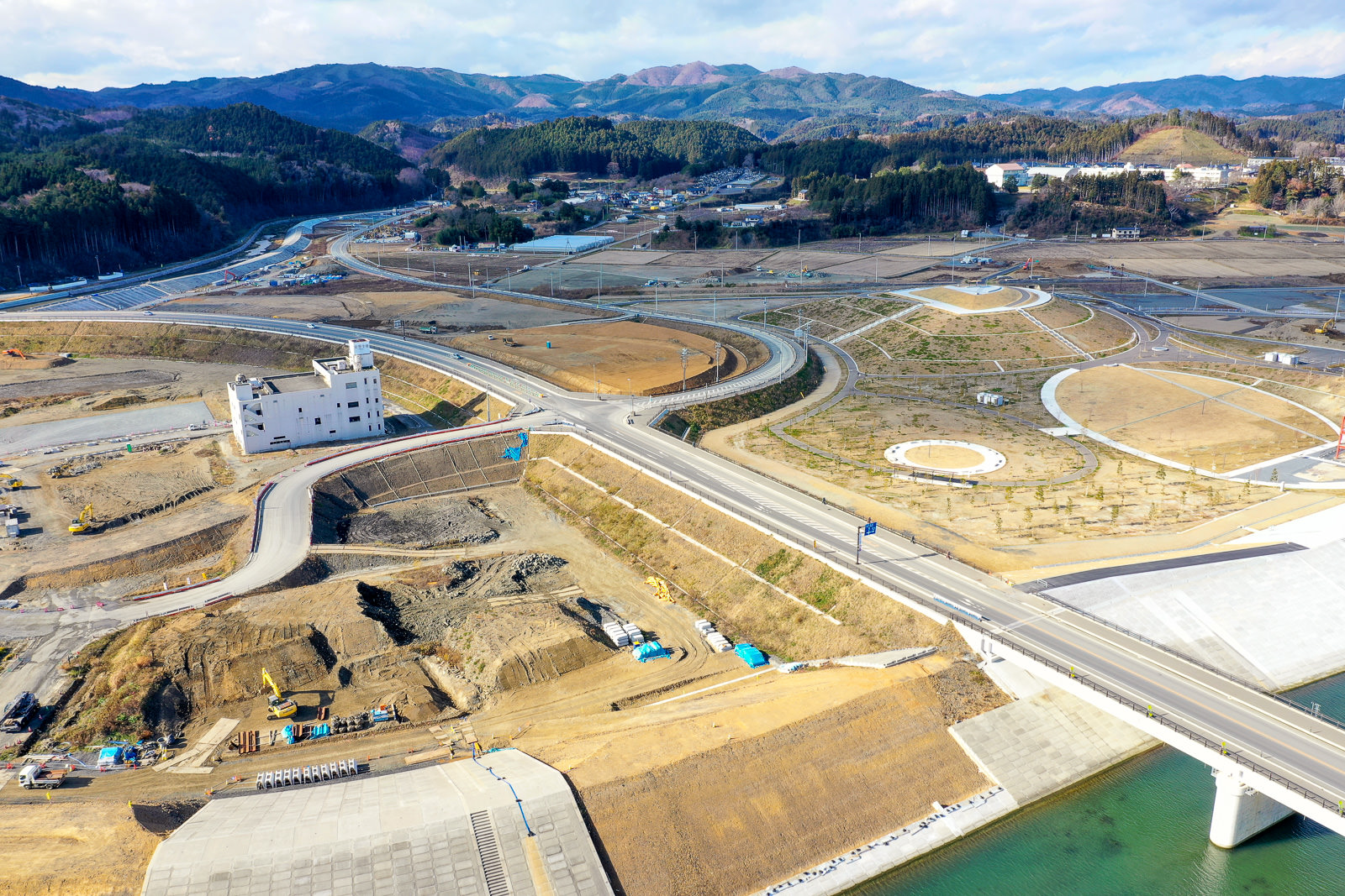 川に架かる橋を中心とした建設現場を上空から撮影した風景で、曲がりくねった道路や小道が敷地内を走り、周囲には丘や山が広がり、木々や草地が点在し、左側には白い建物があり、現場には建設機材や車両が散在する橋と建設現場の写真