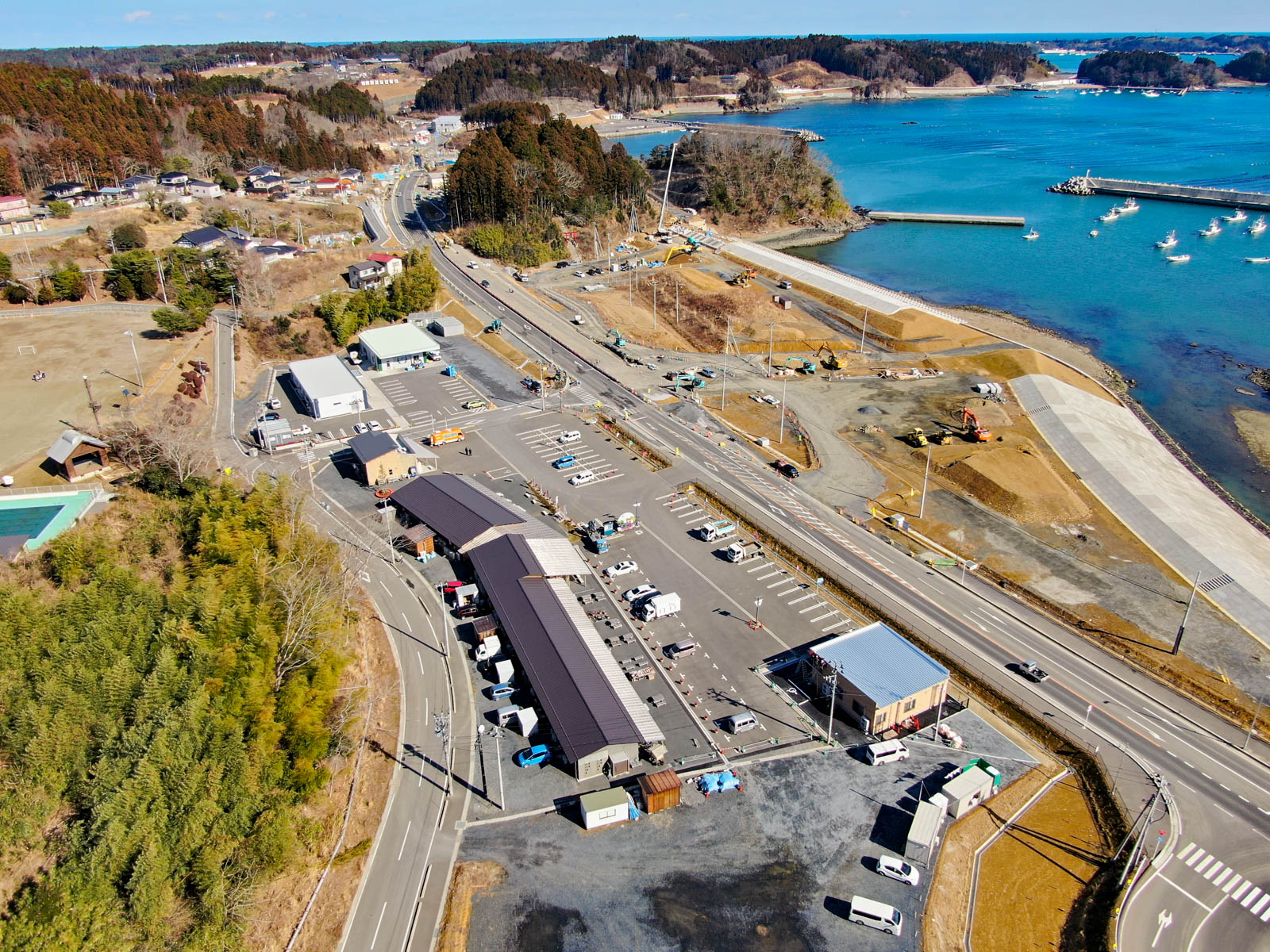 海岸地域を上空から見下ろした風景が広がっており、複数の建物や道路、駐車場が点在し、右側には水域が広がり、桟橋に複数の船が停泊していて、周囲には木々や丘陵地、さまざまな構造物が散在している写真