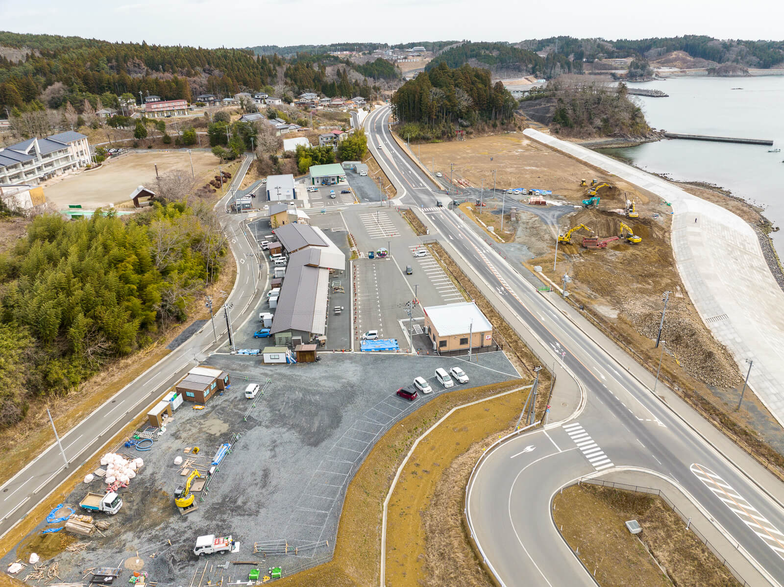 海岸付近に整備された道路と駐車場に囲まれた小規模な建物群が並び、奥に海と防潮堤、工事車両が見える地域の造成中の昼間の風景を高台から定点カメラで撮影した写真