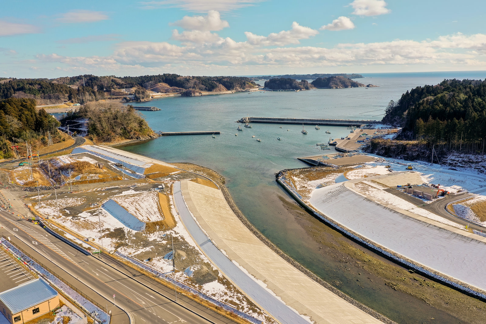 三方を陸地に囲まれた湾を中心に、複数の船が岸近くの海に浮かび、陸地には雪がまだらに積もり、海岸線に沿って主要道路が走り、建物や構造物が海沿いや丘の斜面に点在し、空には雲が少し浮かぶ晴れた冬の日の海辺の湾の写真