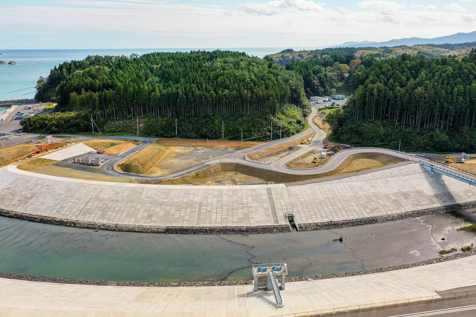 海に面した地域を空から捉えた風景で、手前には津波や洪水対策と思われる大きなコンクリートの堤防があり、その背後には曲がりくねった道路が密集した森林の中を通り、背景には森林に覆われた丘と点在する建物、左側には海、遠くには山々が広がる沿岸防災地域の写真