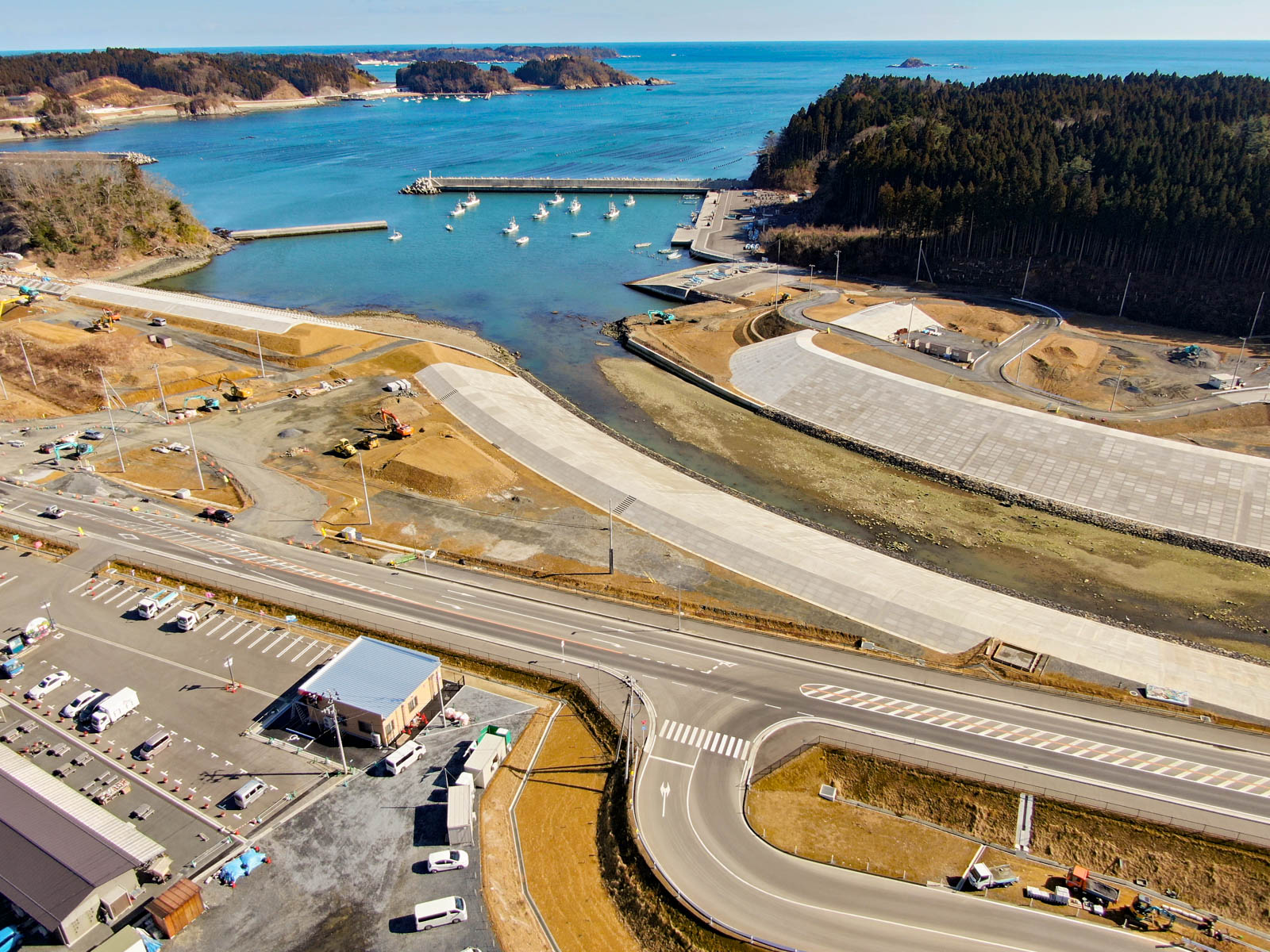 港を中心とした海岸地域を上空から見下ろした風景が広がっており、水面には複数の船が停泊し、海岸線には桟橋や埠頭が並び、周囲には道路や駐車場、建物が点在し、一方には森林地帯が広がっていて、港全体は整備された海洋活動のためのインフラが整っており、水面は穏やかで混雑の様子は見られない写真