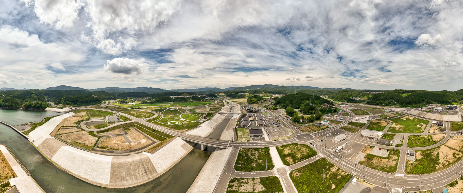 中央にまっすぐ伸びた一本の川が流れ、左右に広大な土地が見え、舗装された道路や橋が整備されている地域全体の昼間の風景を高台から定点カメラで撮影した写真