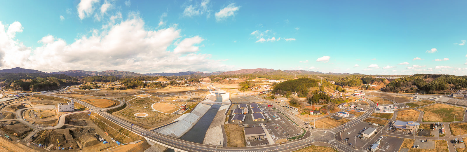 自然の風景と開発されたインフラが混在する農村地域を広く見渡した風景で、道路や建物、大規模な建設現場のような場所があり、背景には山々と森林が広がり、空には雲が点在する農村地域の写真