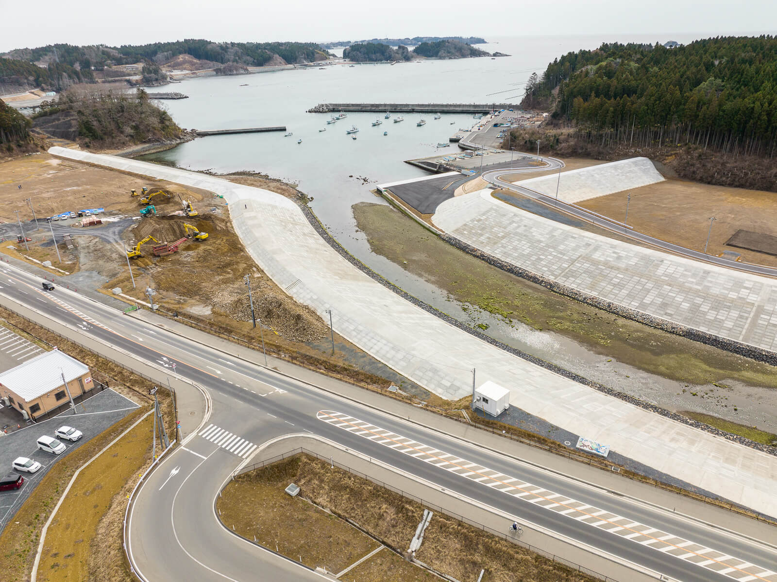 護岸工事が施された川が海へと流れ込み、湾内に停泊する漁船とそれを囲む堤防、道路が湾曲しながら海へ続く様子を示す港湾周辺の昼間の風景を高台から定点カメラで撮影した写真