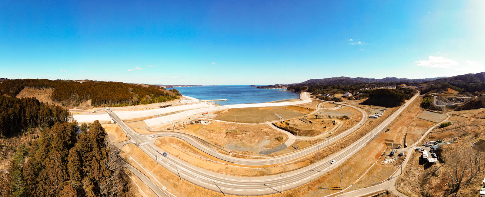 海岸地域のパノラマ風景が広がっており、曲がりくねった道路に車両が走っていて、周囲には丘陵地が広がり、背景には穏やかな海が見え、空は雲がほとんどない澄んだ青空が広がっている写真
