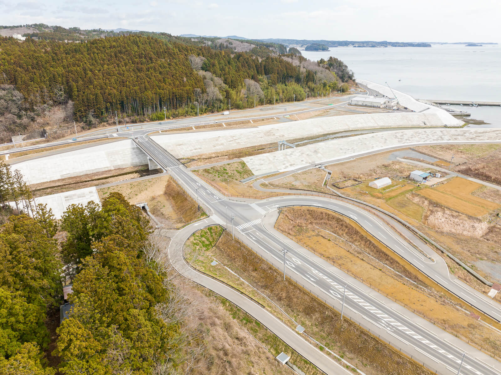 山林を背にして、防潮堤に沿って新しく建設された道路と橋が交差し、周囲には農地と小さな建物が点在している、海辺に近いインフラ整備の昼間の風景を高台から定点カメラで撮影した写真
