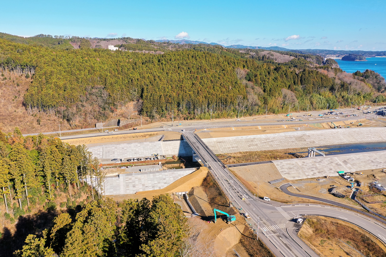 森に囲まれた丘陵地帯の中にある建設現場を上空から撮影した風景で、現場には複数の大きなコンクリート板が並べられ、橋がその上を横断しており、遠くには水辺が見え、建設機材も点在している建設現場の写真