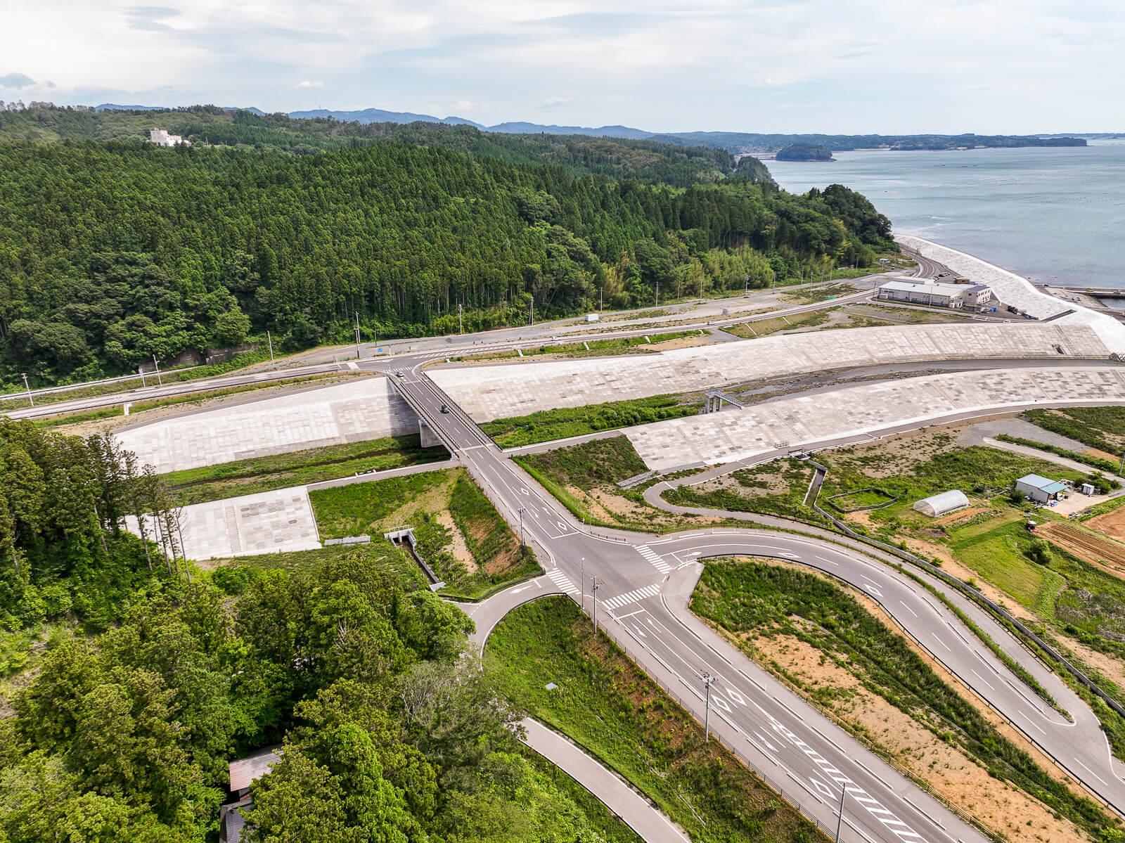 海沿いに大規模な防潮堤が築かれ、それに沿って道路が整備され、森と海の間にある交差点が見える昼間の風景を高台から定点カメラで撮影した写真