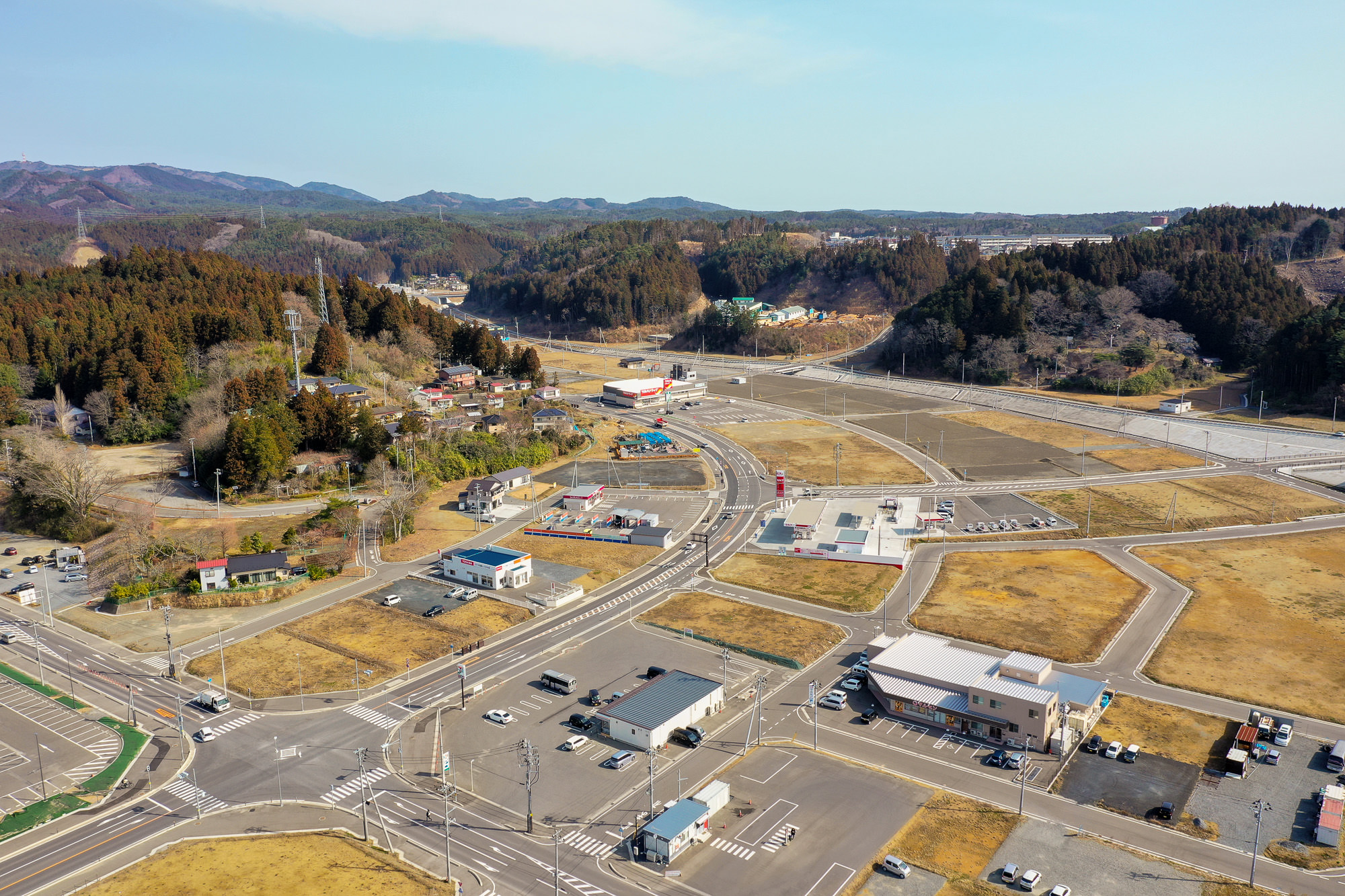 建物や商業施設が点在する田舎の地域を空から見下ろした写真で、道路が格子状に交差し、周囲には木々が密集した丘が広がっている写真