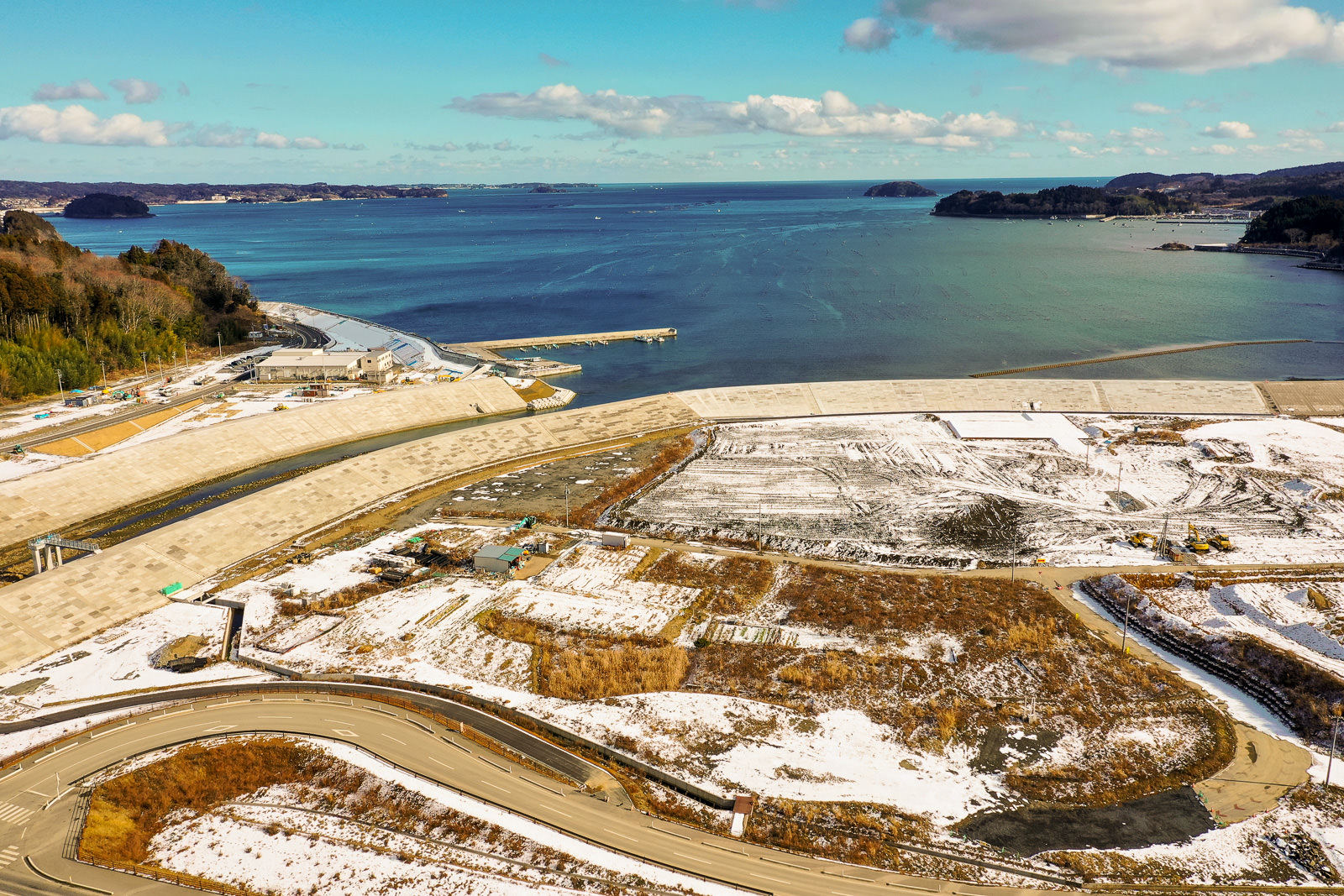 雪が部分的に積もった自然と開発地が混在する海岸地域を上空から撮影した風景で、手前には曲がりくねった道路が走り、周囲には草地や雪に覆われた土地が広がり、奥には造成地または畑のような広い平地があり、その先には水面と複数の小島が見え、空は晴れて雲が少し浮かぶ冬の海岸地域の写真