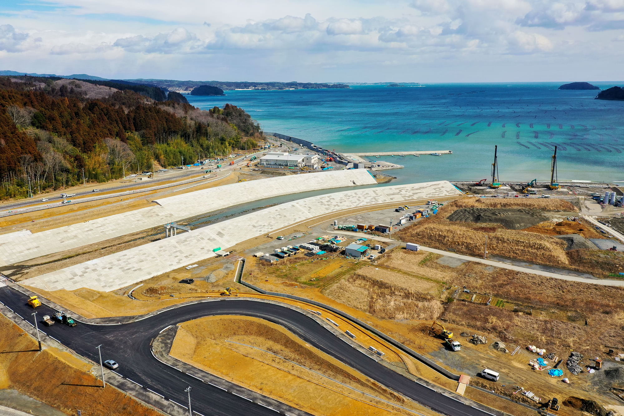 海岸沿いの工事現場を空から撮影した写真で、手前には舗装された道路が伸び、奥には防波堤や建設機械が配置されている様子が見える写真