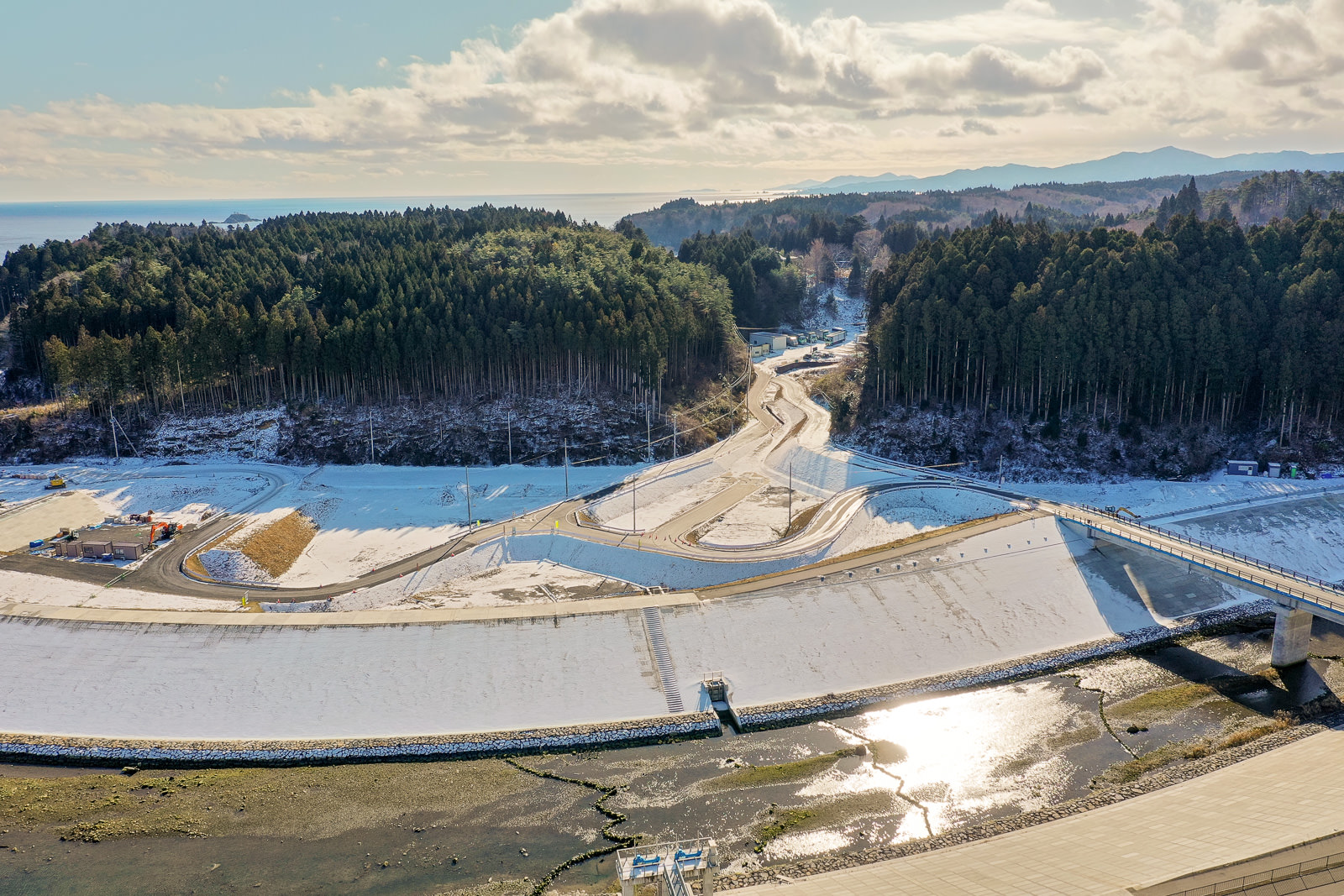 雪に覆われた曲がりくねった道路が通り抜け、両側には密集した森林が広がり、遠くには山々が連なり、空には雲が点在する冬の風景の写真