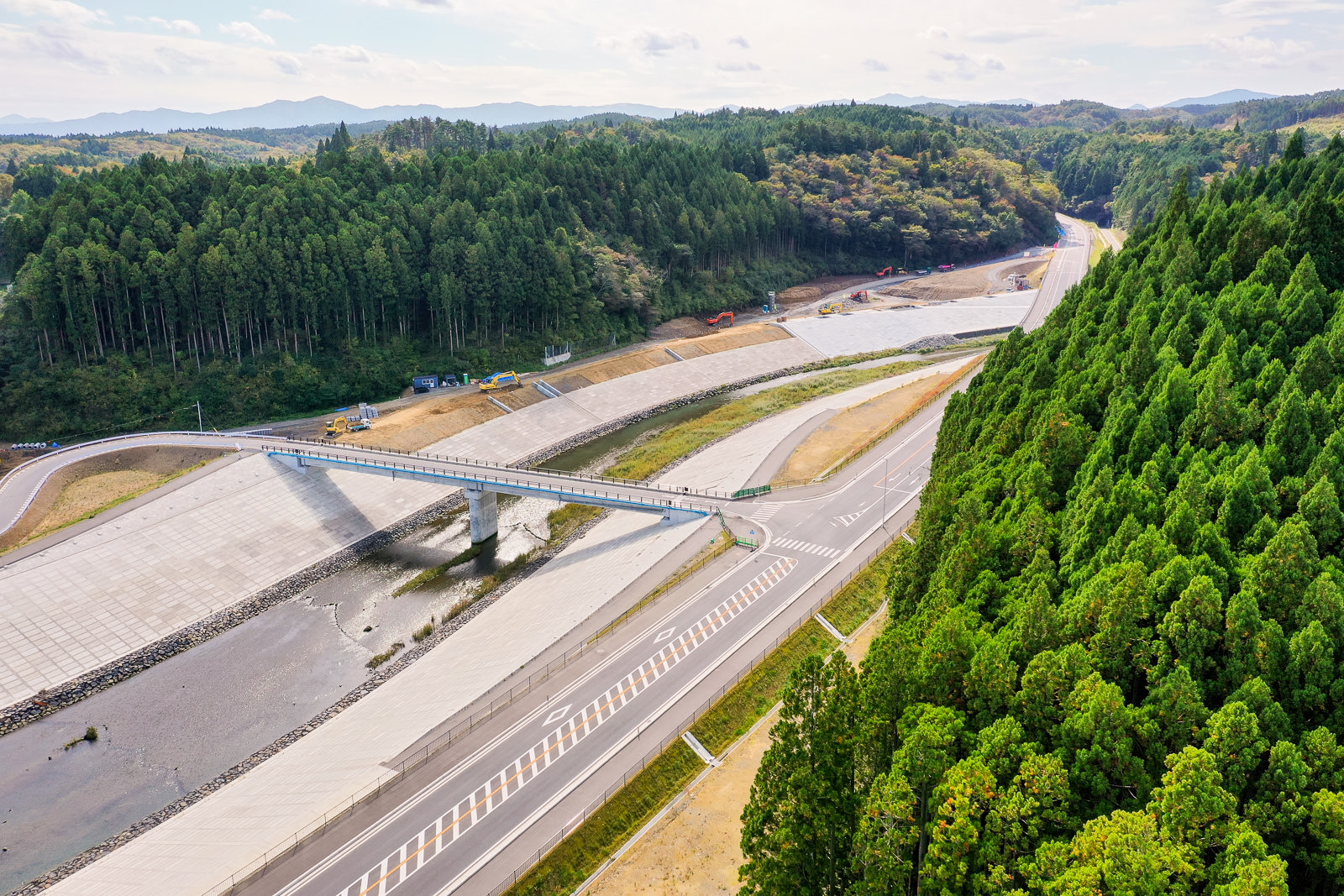 川に架かる橋を中心に、周囲を密集した森林と丘陵が囲み、橋は複数車線の道路網の一部として整備されている自然と交通インフラが融合した地域の写真
