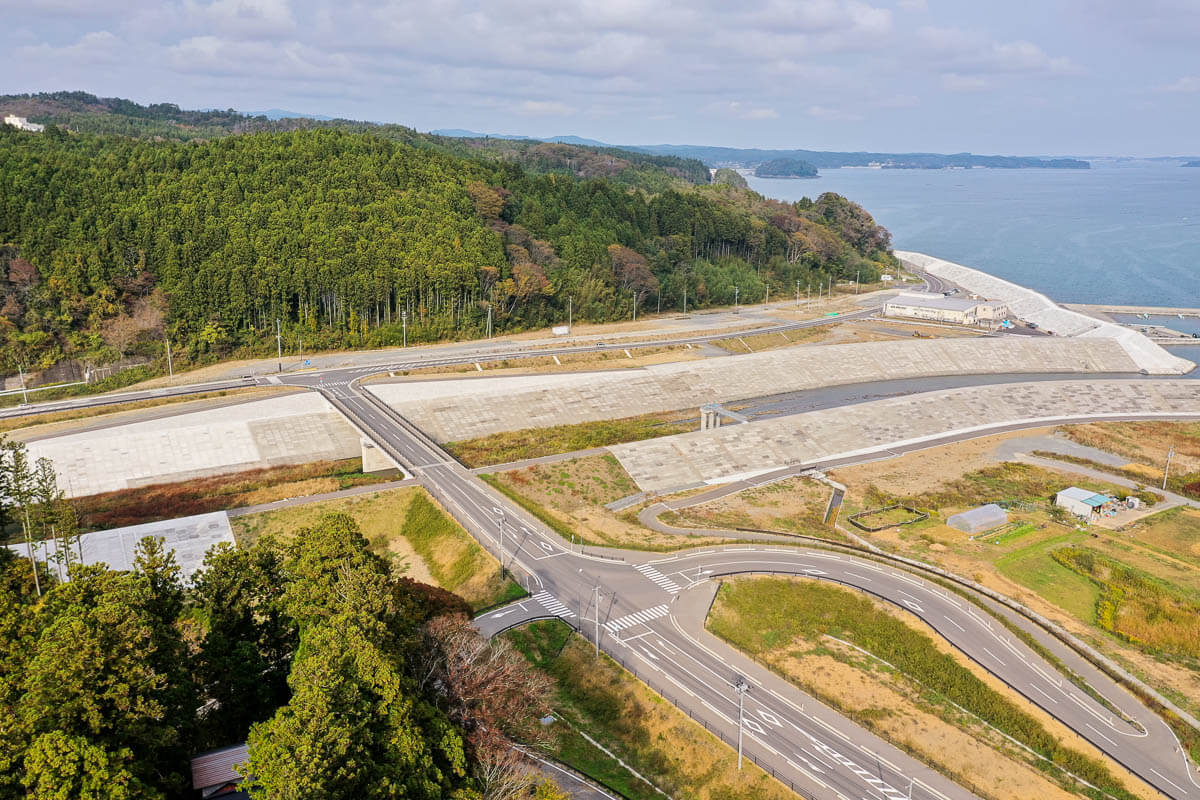 緑豊かな山と青い海を背景に、防潮堤と直線的な道路や交差点が整備された沿岸部の風景の写真