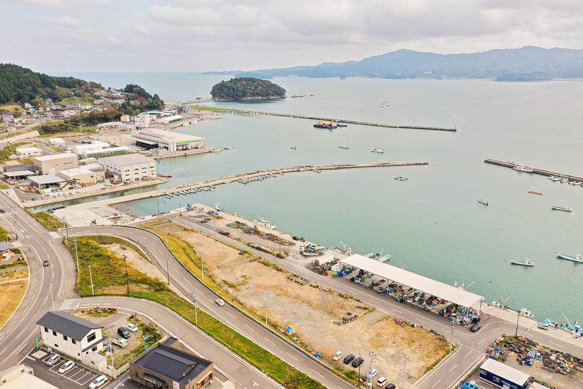 港町の道路沿いに並ぶ建物や市場施設、その先に広がる防波堤と停泊する漁船、背景に小島と山並みが見える海辺の風景の写真