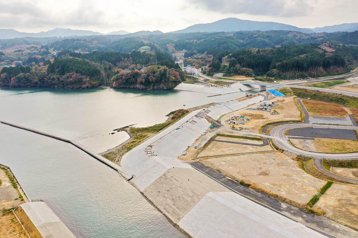 海に面した防潮堤と造成地、工事用車両が並ぶ土地、その奥に橋と道路、紅葉を帯びた山並みと入り江が広がる風景の写真