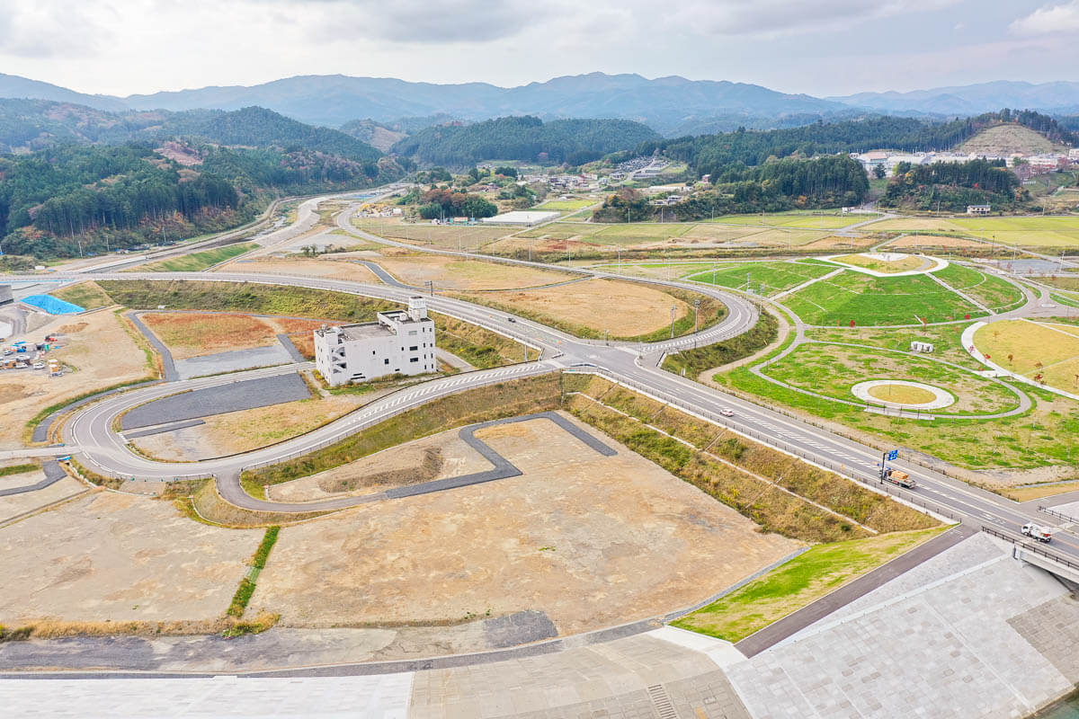 造成地の中央に白い建物が残り、周囲には曲線的に整備された道路と円形の緑地広場、背後に田畑と山並みが広がる風景の写真