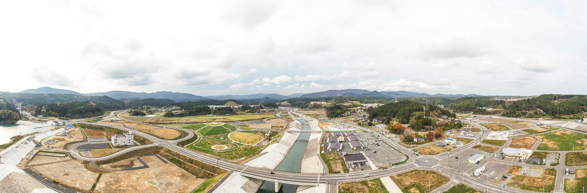 河川と堤防を中心に造成地や整備された道路網、円形の緑地広場、駐車場や建物群が広がり、背後に山々と町並みが見えるパノラマ風景の写真