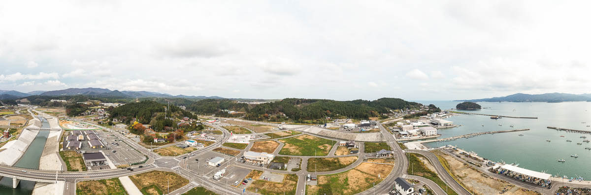 河川と堤防沿いに広がる整備された街並みと駐車場、さらに港湾施設や停泊する船が見える海、背後に山並みが広がるパノラマ風景の写真