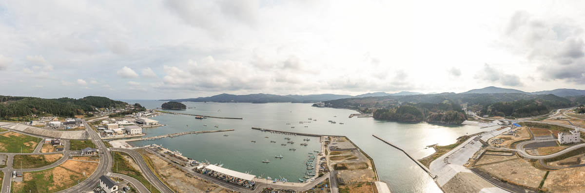 広い湾内に多くの漁船が停泊し、防波堤や港湾施設が整備された海辺の町並みと、背後に山並みが広がるパノラマ風景の写真