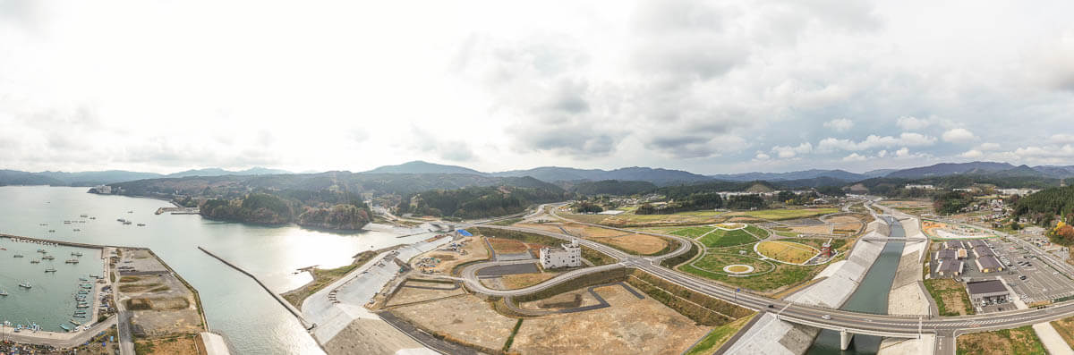 港に停泊する漁船や防波堤、造成地と河川、整備された緑地公園や道路網、背後に山並みが広がる沿岸部のパノラマ風景の写真