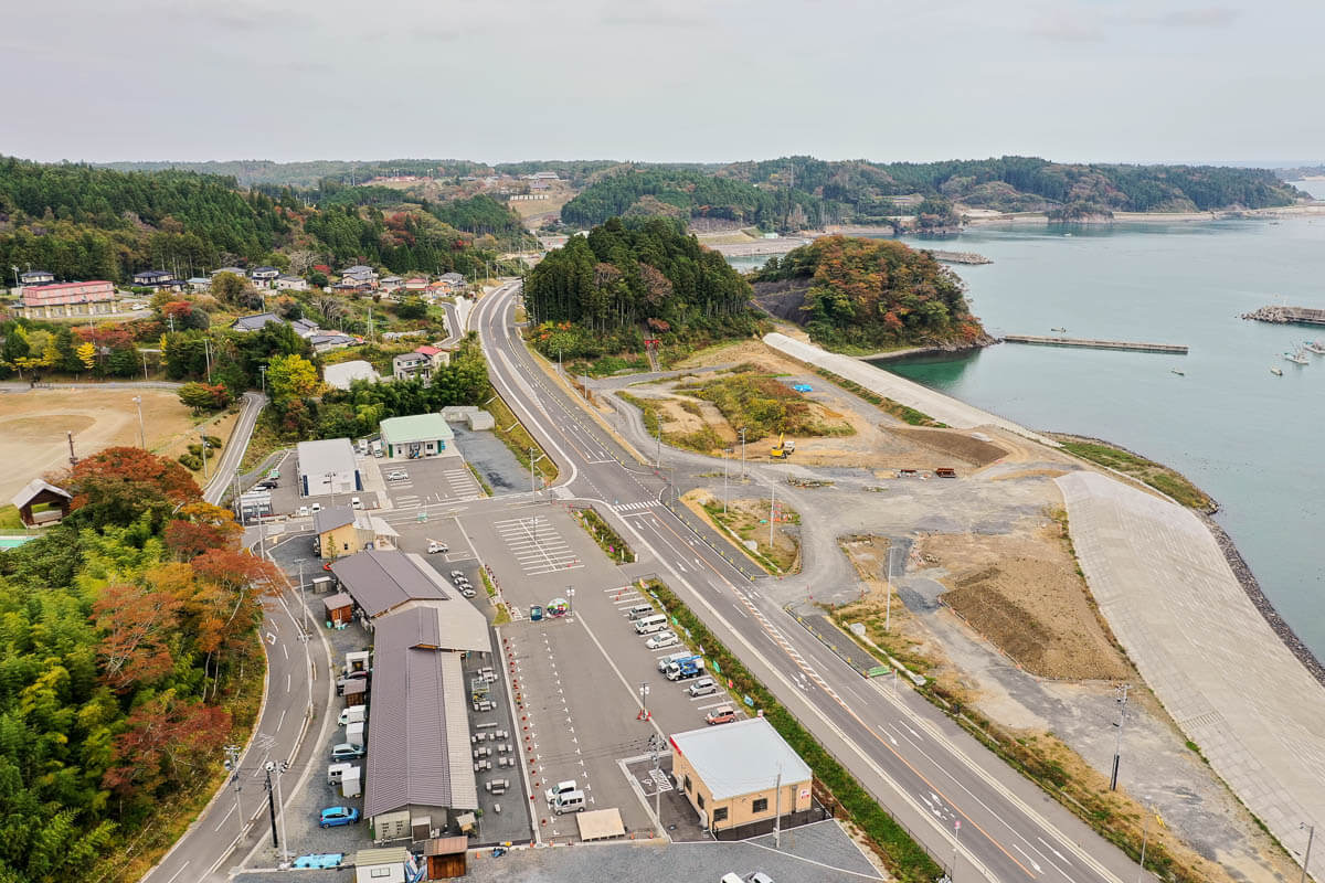 道路沿いに整備された駐車場と建物群、その先に小さな岬と防潮堤、湾内に浮かぶ漁船や背後の緑豊かな山々が広がる海辺の風景の写真
