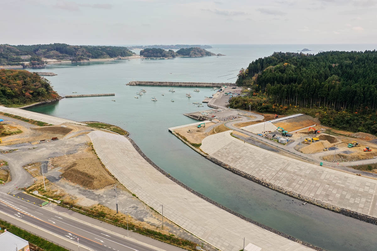 防潮堤に囲まれた湾内に漁船が浮かび、港湾施設や工事中の造成地、周囲を覆う森林と小島が広がる沿岸部の写真