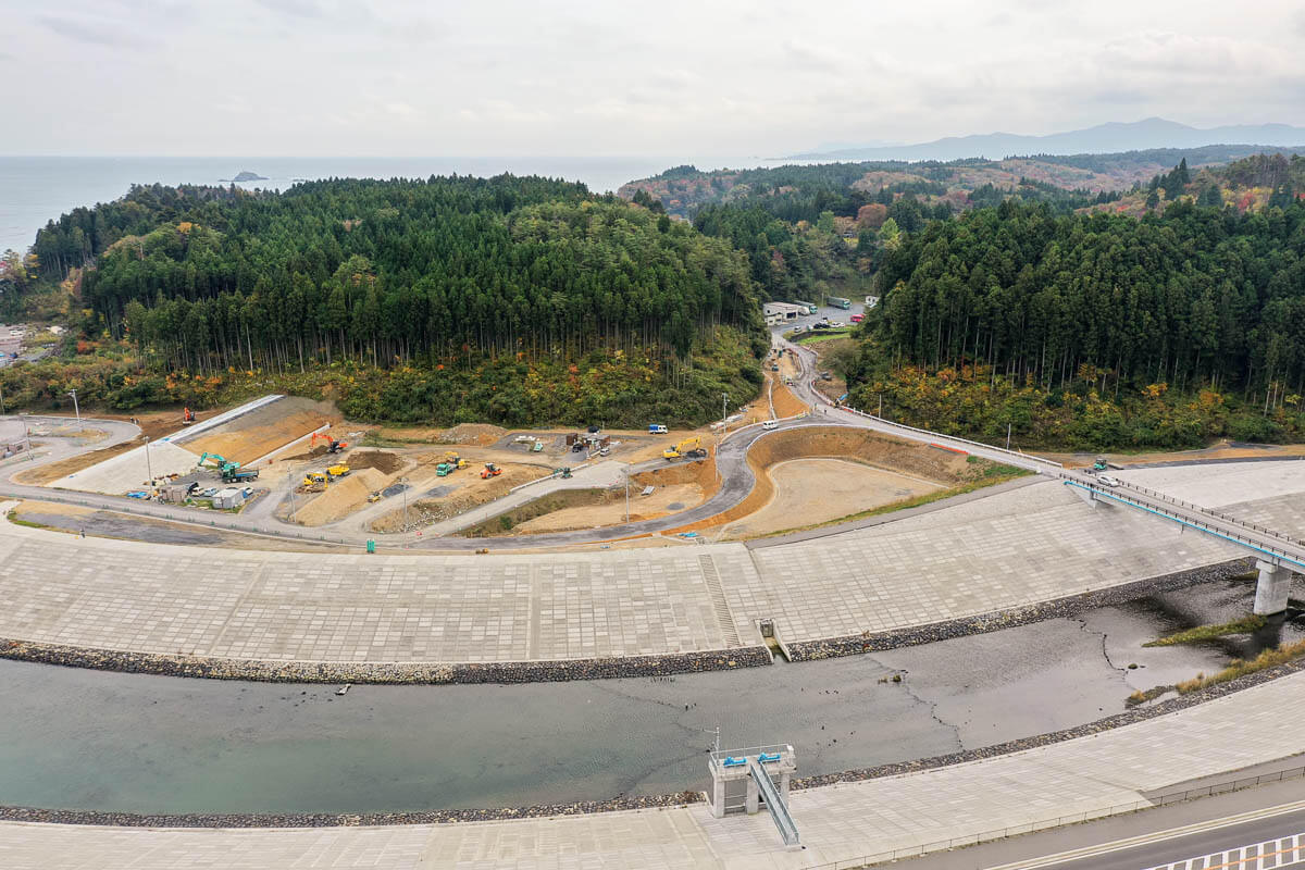 護岸に囲まれた河口部と、奥に広がる森と造成工事現場を捉えた写真。重機が作業する造成地と舗装された道路が見え、背後には針葉樹林と山並みが広がっている写真
