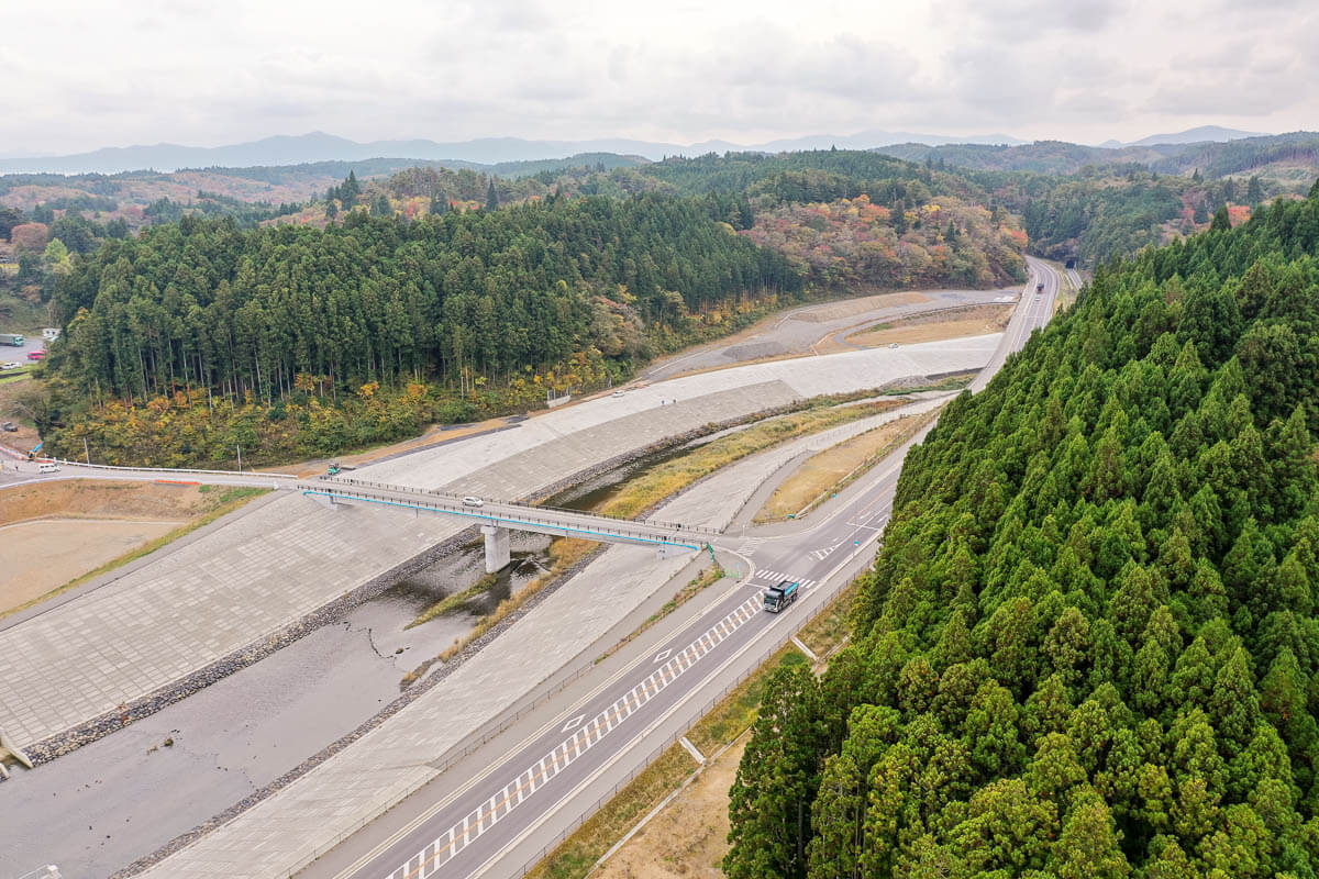 手前には走行中のトラックが見え、奥には舗装道路が山間部へと続いて両側には杉林を中心とした濃い緑の森が広がり、秋の紅葉がところどころ彩りを添えている堤防に沿った川と、その上に架かる橋を捉えた写真