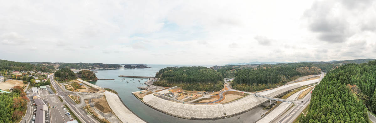 防潮堤に囲まれた湾と漁船の浮かぶ港、緑豊かな小高い山や造成地、橋と道路が調和する沿岸部のパノラマ風景の写真