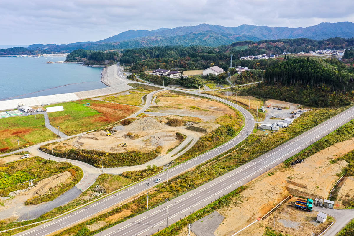 海沿いに築かれた防潮堤とカーブを描く道路、造成中の土地や建設機械、背後に山並みと集落が広がる風景の写真