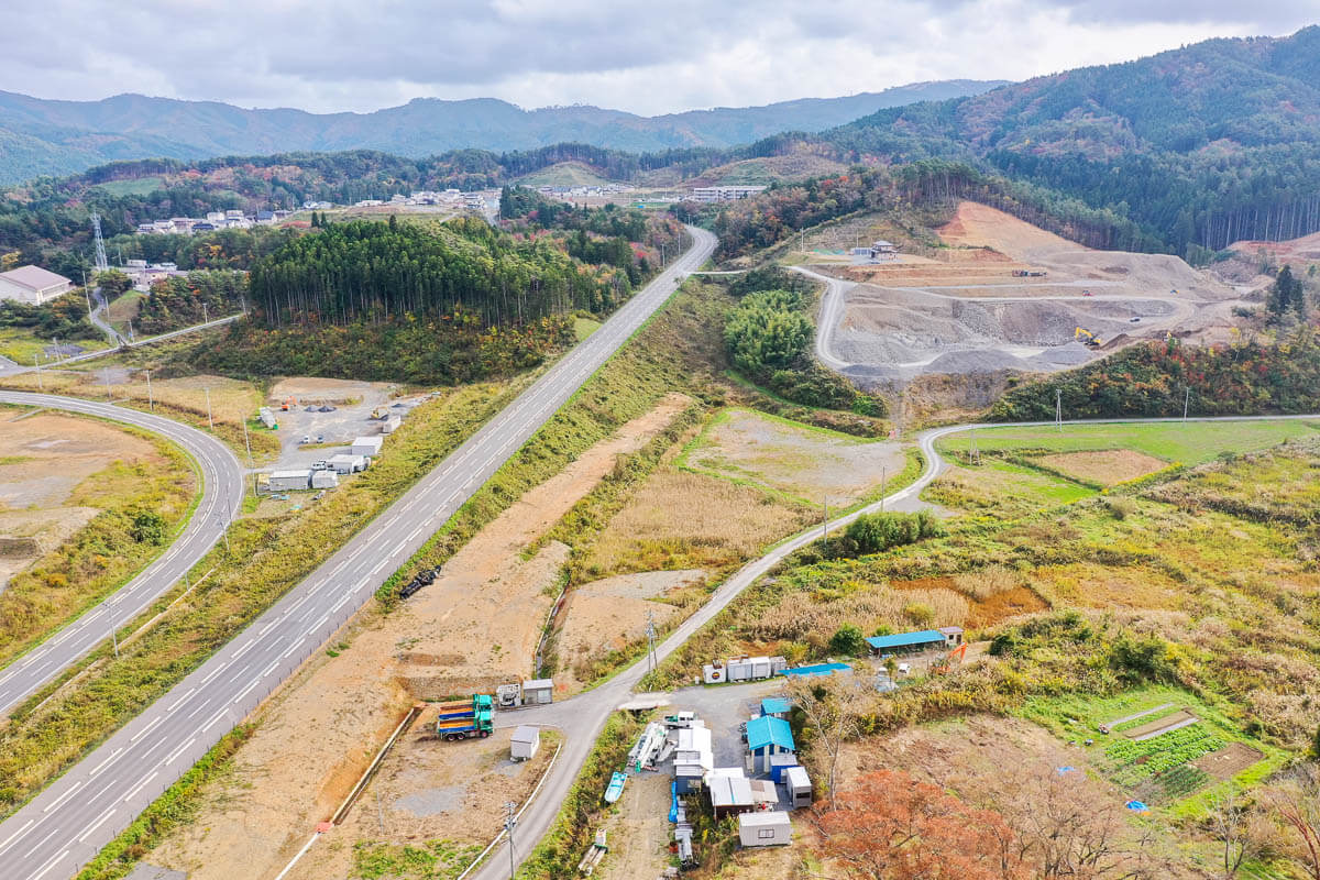 山間に延びる道路と造成工事中の広大な土地、点在する住宅や畑、背後に紅葉を帯びた山並みが広がる風景の写真