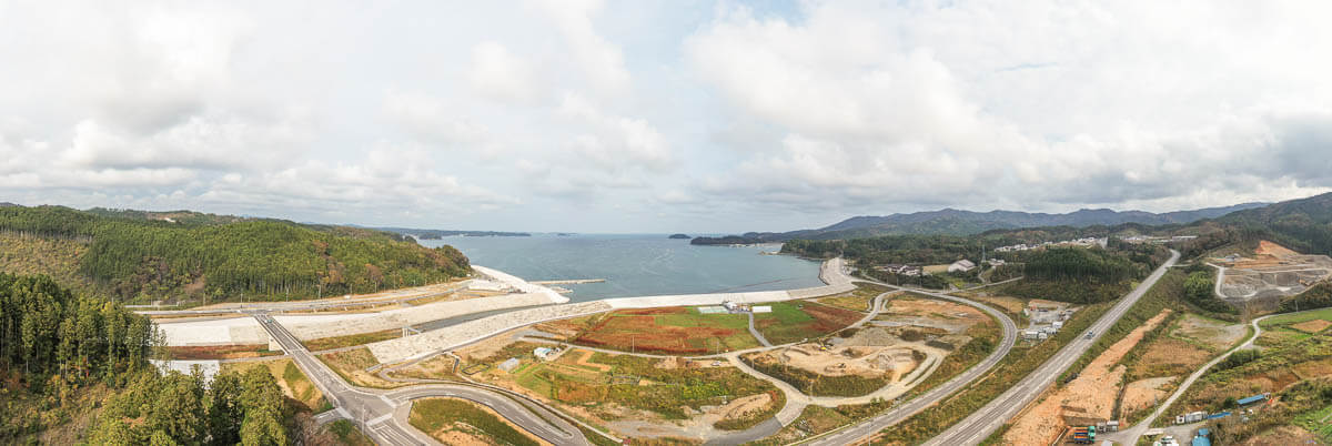 海に面した湾岸と防潮堤を中心に、緑の山々や造成地、曲がりくねる道路が広がる沿岸部の景観の写真