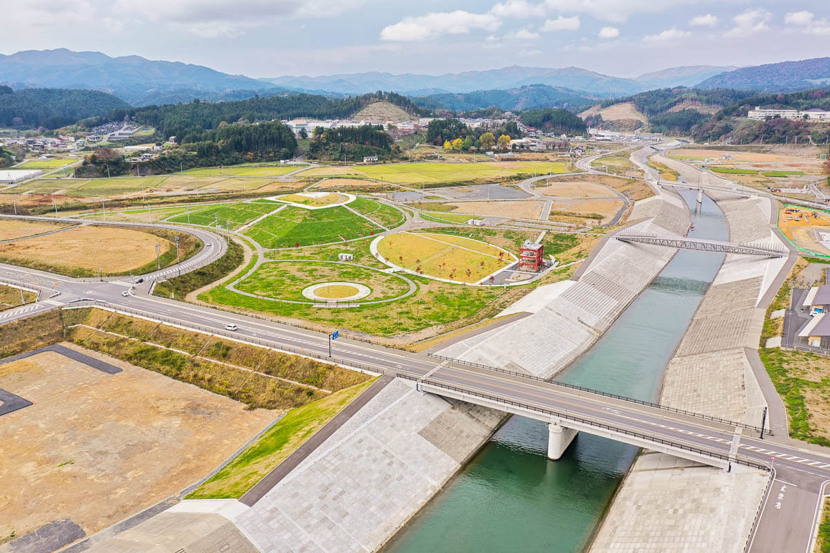 堤防に囲まれた河川沿いに整備された緑地公園と円形の広場、道路や橋梁が整然と配置され、奥に田畑と山々が広がる風景の写真