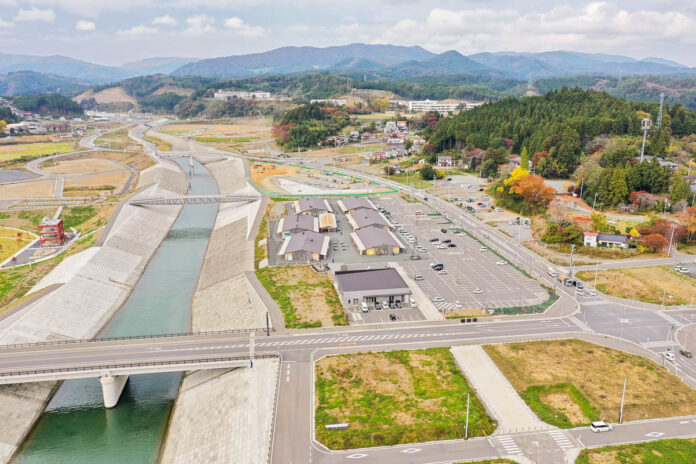 山々を背景に広がる町並みと広大な駐車場を持つ施設群、中央を流れる川と堤防に整備された橋梁が見える風景の写真