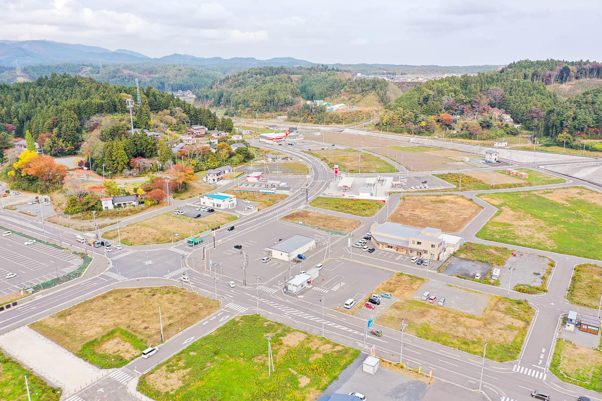 交差点を中心に整備された道路と駐車場、コンビニやガソリンスタンドが並ぶ街並み、背後に森林と山々が広がる風景の写真