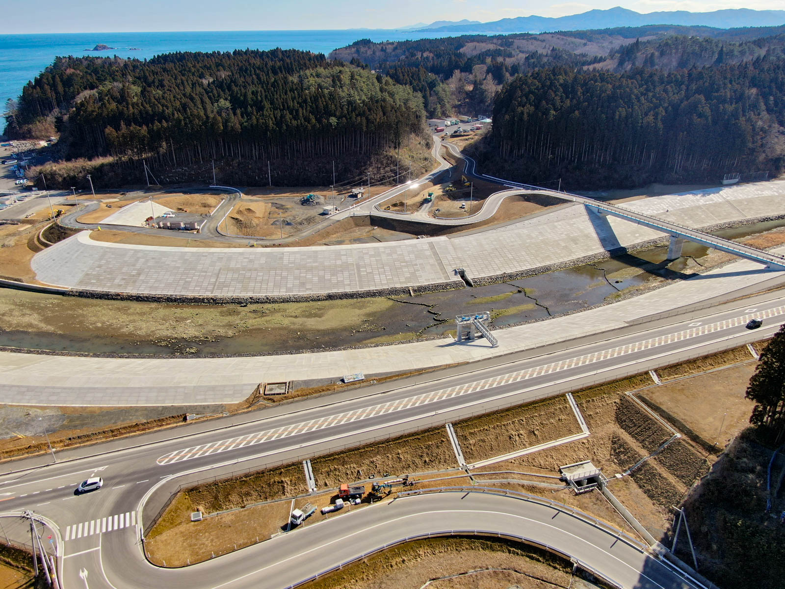 海岸地域を上空から見下ろした風景が広がっており、複数の曲がりくねった道路と大きなコンクリート製の堤防があり、その上を橋が横断していて、周囲には密集した森林と丘陵地が広がり、背景には海が見える写真