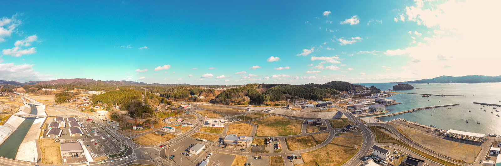 海沿いに位置する町を上空から広く見渡した風景で、町にはさまざまな建物や道路、インフラが整備されており、海岸には桟橋や波止場が突き出し、背景には丘や森林が広がり、空には雲が点在する沿岸の町の写真