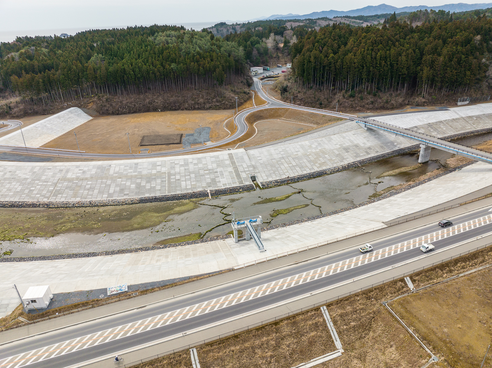 大きな護岸コンクリートで囲まれた川にかかる橋と、川沿いの道路や山へと続く道が見え、全体に護岸工事が施されている昼間の風景を高台から定点カメラで撮影した写真
