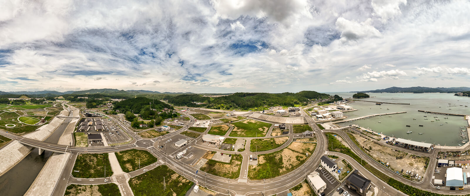 港と道路、建物が広がる町の遠景が見える川沿いの地域をパノラマで捉えた昼間の風景を高台から定点カメラで撮影した写真