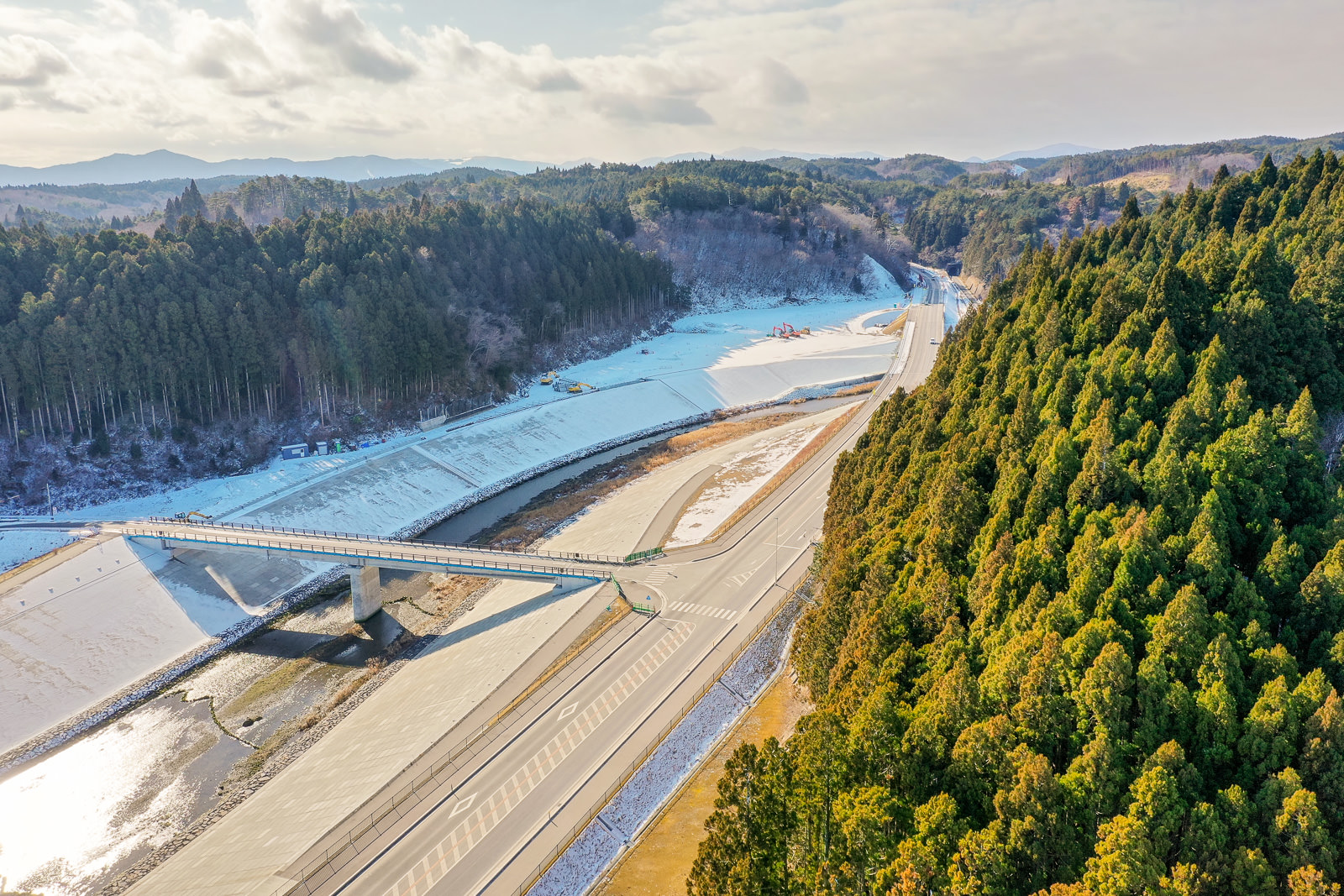 森林に囲まれた地域を上空から撮影したもので、道路が中央を通り、その上に橋が架かっており、周囲には密集した森と丘が広がり、地面には雪がまだらに積もり、空には雲が点在しながらも日差しが差し込む冬の道路の写真