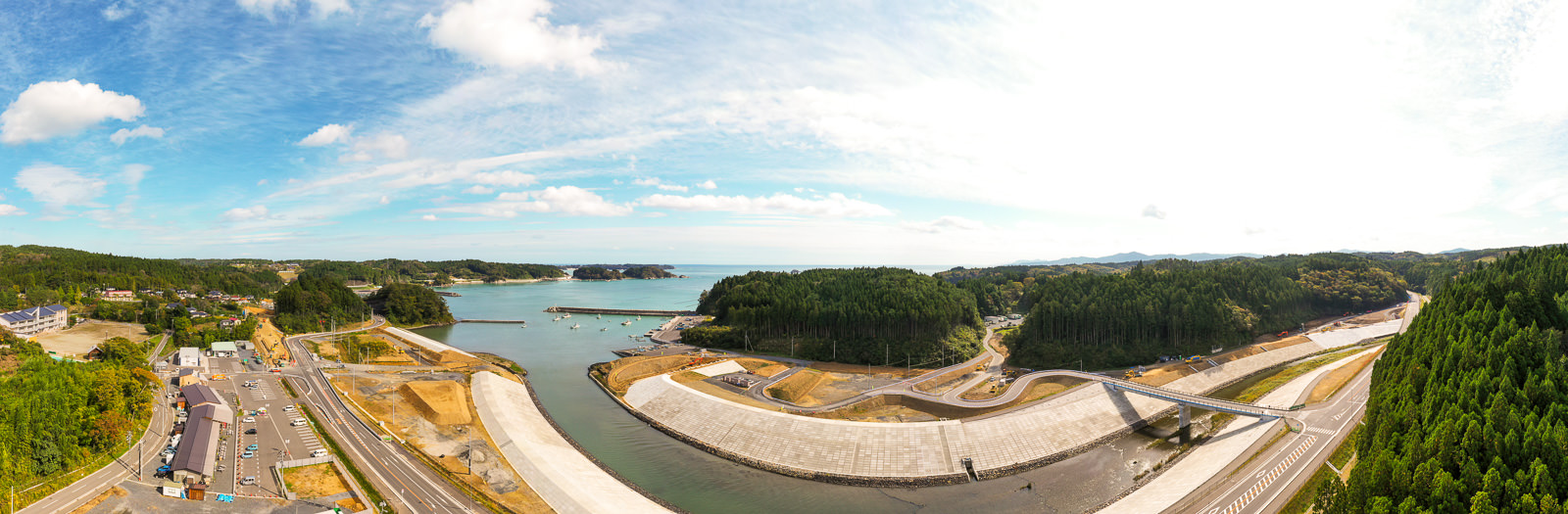 中央に水域が広がり、それを横断する大きなコンクリートの防災堤防が目立ち、周囲には緑豊かな森林と丘陵が広がり、左側には道路や建物が点在する人の暮らしと自然が共存する沿岸地域の写真
