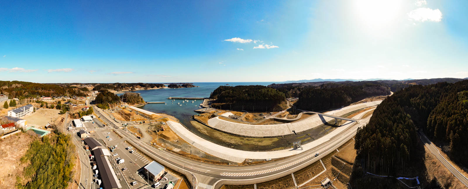 大きな湾または入り江を中心とした海岸地域のパノラマ風景が広がっており、手前には道路や建物、ダムまたは防潮堤のようなインフラが見られ、背景には森林に覆われた丘と広がる海があり、空は青く澄み、ところどころに雲が浮かぶ写真