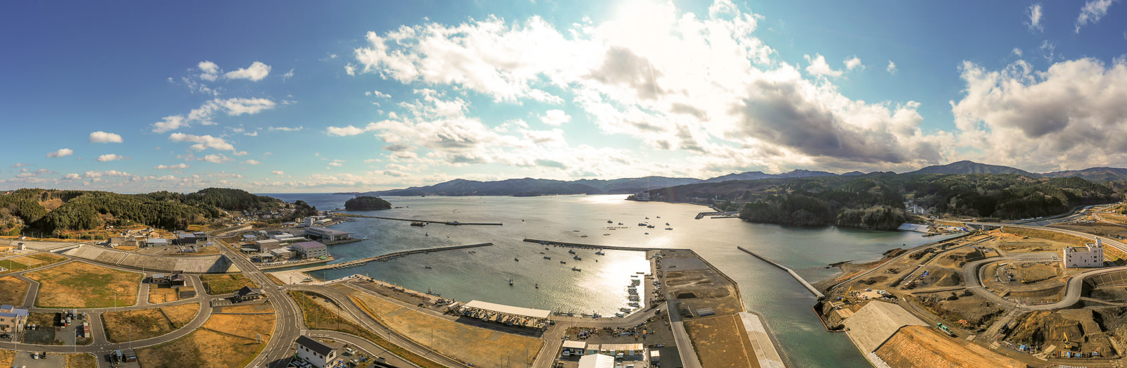 港を中心とした海岸地域を広く見渡した風景で、海には複数の船が浮かび、海岸線は丘や山に囲まれており、空には雲が点在しながらも日差しが水面に反射し、港周辺には道路や建物、建設中の場所も見られる沿岸の港町の写真