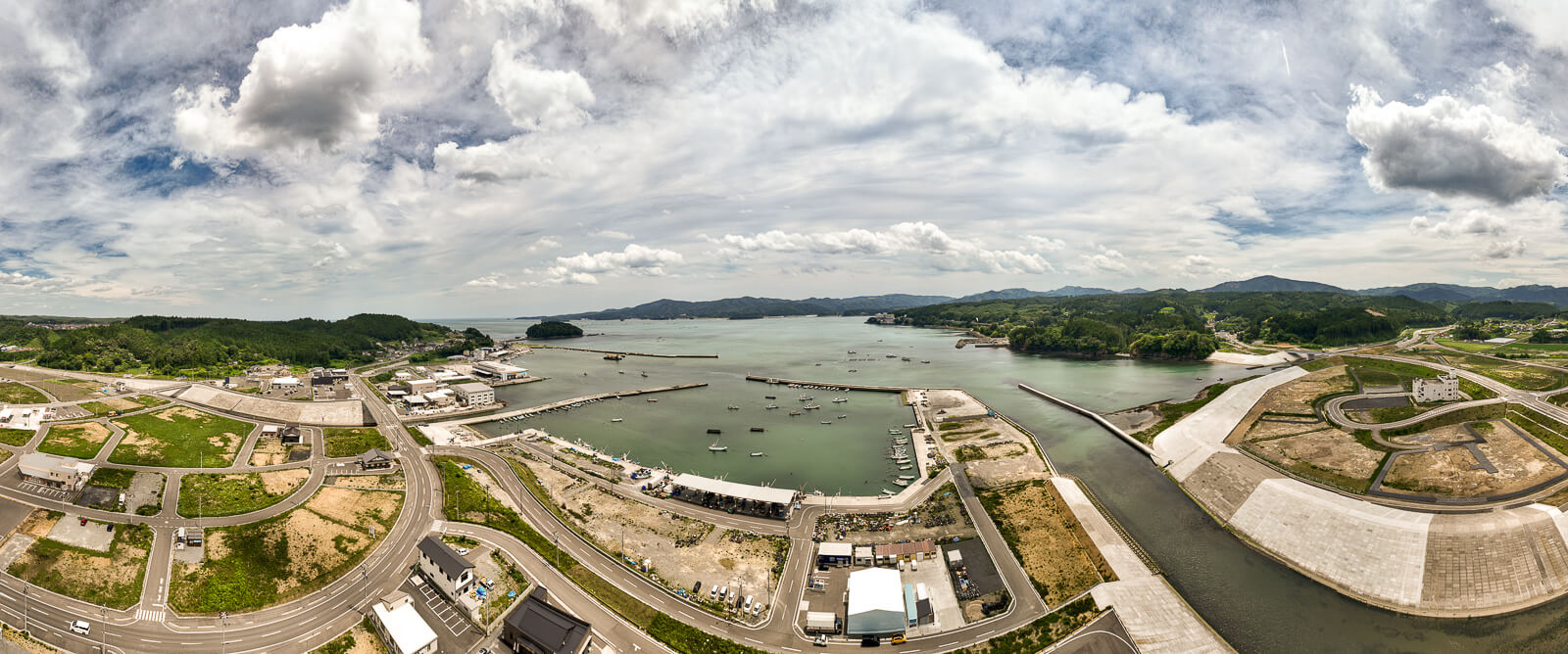 広範囲の湾と港、周囲に道路や建物が点在し、遠くに山々が見えるパノラマ景観の昼間の風景を高台から定点カメラで撮影した写真