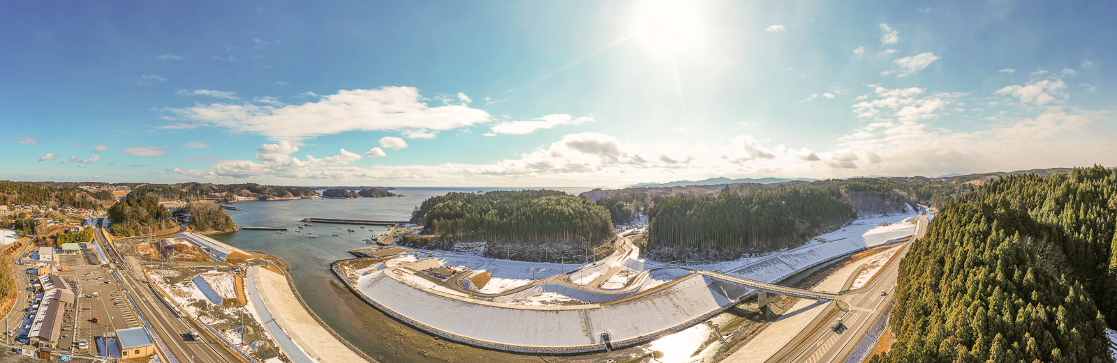 入り江または湾と思われる水域を中心に、周囲には森林に覆われた丘と雪が積もった地面が広がり、左側には道路が走り、建物や車両が点在し、空は雲が少し浮かぶ中で太陽が明るく輝いている冬の海辺の風景の写真