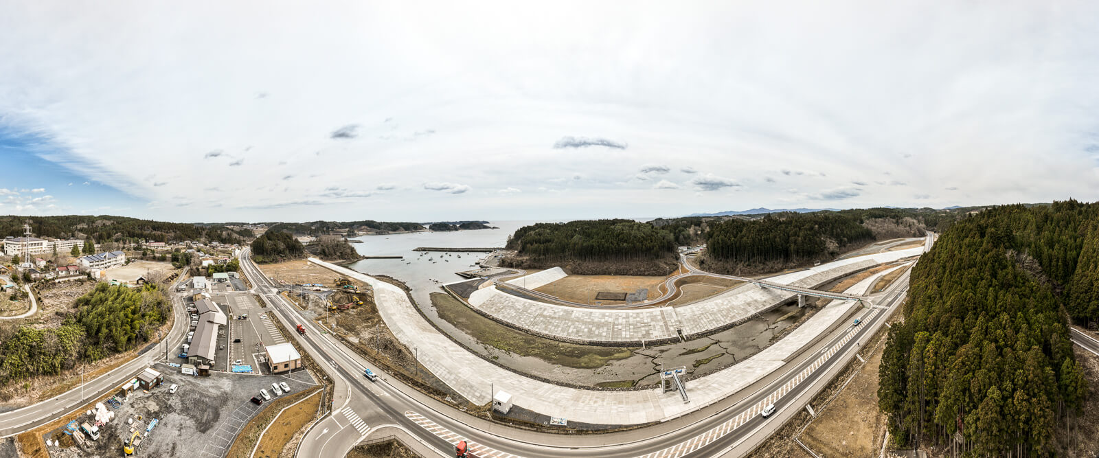 海岸線から山に囲まれた地域まで広く見渡せる、防潮堤と川の護岸整備が進む湾に面した小さな町の昼間の風景を高台から定点カメラで撮影した写真