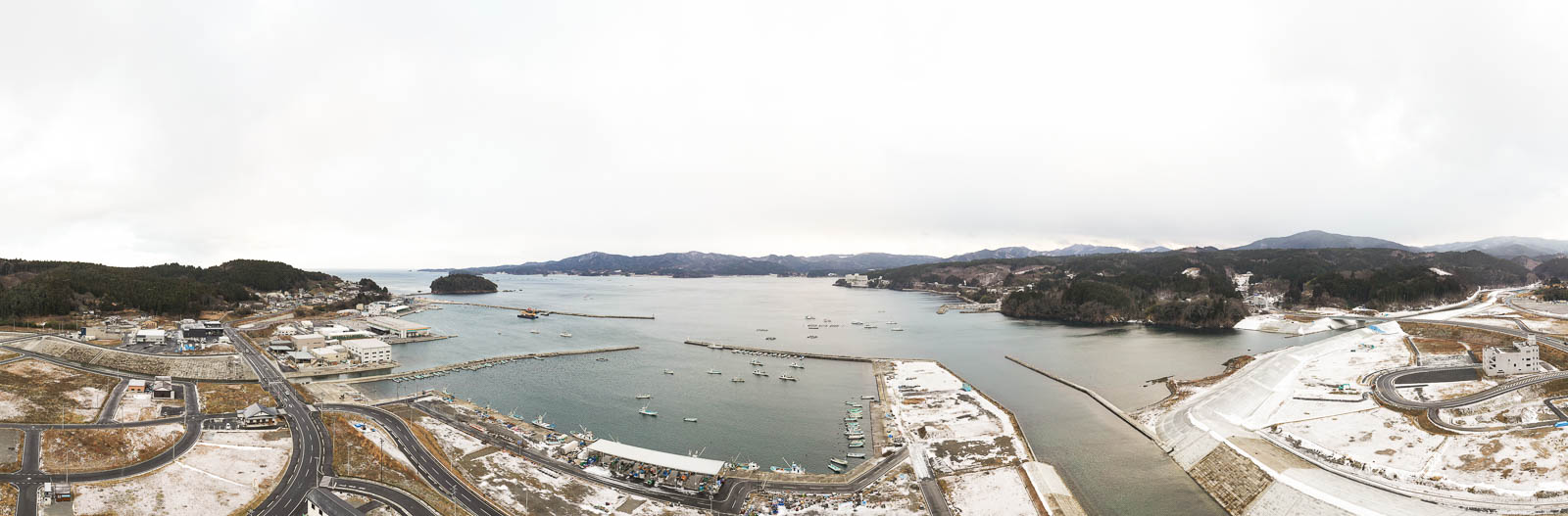 湾に沿って広がる港町と漁船が並ぶ海、奥には防波堤と山々が広がるパノラマの昼間の風景を高台から定点カメラで撮影した写真